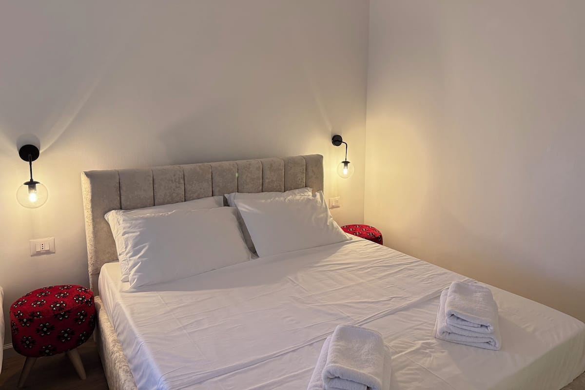 Ein ordentlich bezogenes Bett mit weißen Leinen, zwei Nachttischlampen und ein rot gemustertes Ottoman.