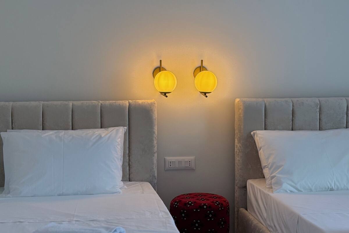 Ein Hotelzimmer mit zwei Betten, jeweils mit grauem Kopfteil und weißer Bettwäsche. Zwei Wandleuchten befinden sich über den Betten.