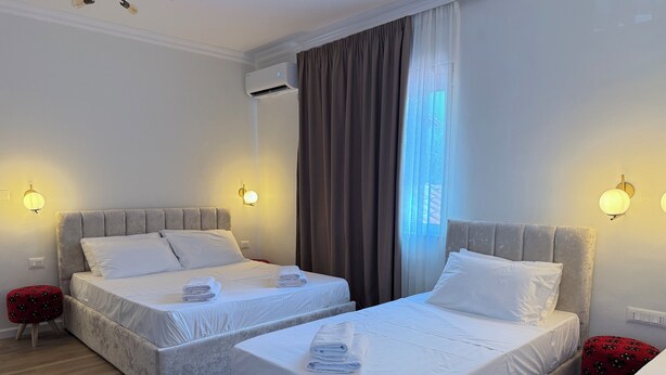 Ein Hotelzimmer mit zwei Betten, einem roten Hocker, einer Wand-Klimaanlage und einem Fenster mit Vorhängen.