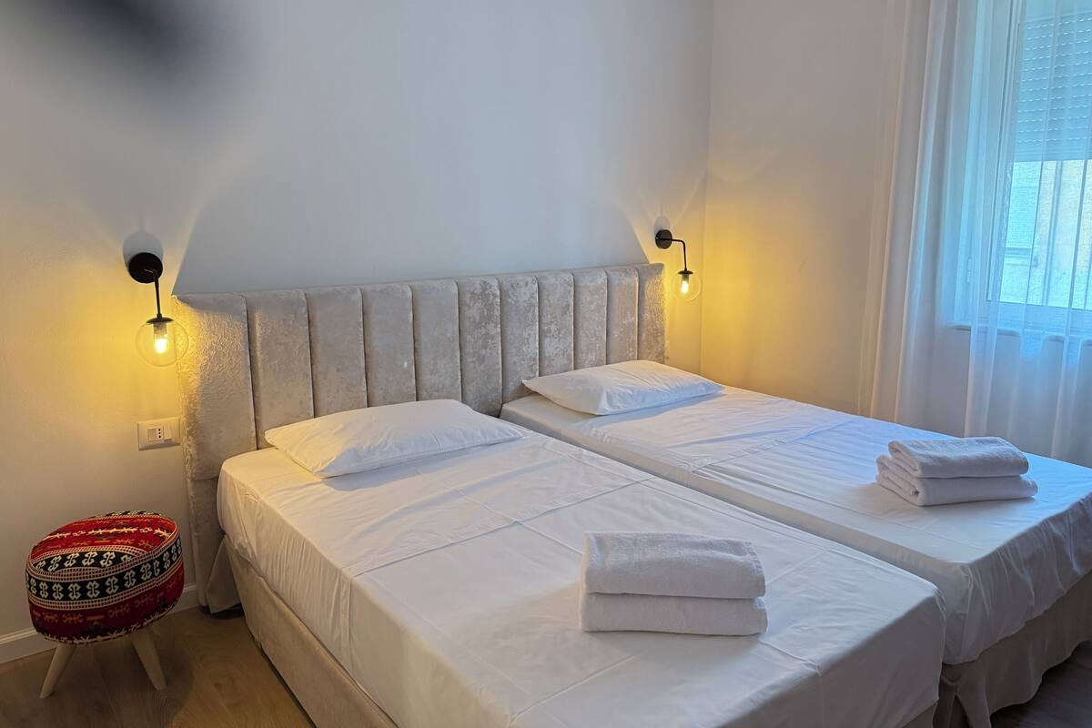 Ein Hotelzimmer mit zwei Betten, einem kleinen Hocker und einem Fenster mit Vorhängen.