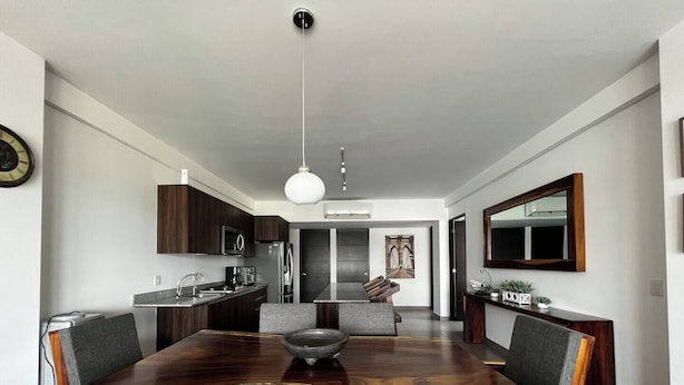 A modern kitchen with dark wood cabinets, a central island, and a dining area with a large wooden table and chairs. - Parota Apartment (Nuevo Vallarta)