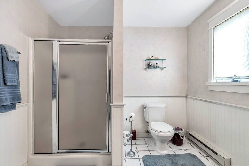 Une salle de bain avec une douche à parois de verre, des toilettes, une petite étagère avec des articles de toilette et une fenêtre avec des stores.
