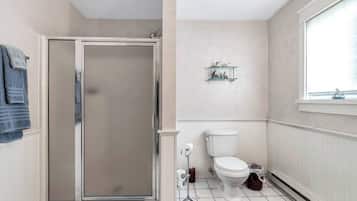 Une salle de bain avec une douche à parois de verre, des toilettes, une petite étagère avec des articles de toilette et une fenêtre avec des stores.