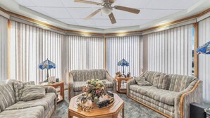 A corner living room with striped curtains, a ceiling fan, two sofas, a coffee table with a floral centerpiece, and two lamps. - Red Barn Village Apartments (Clarks Summit)