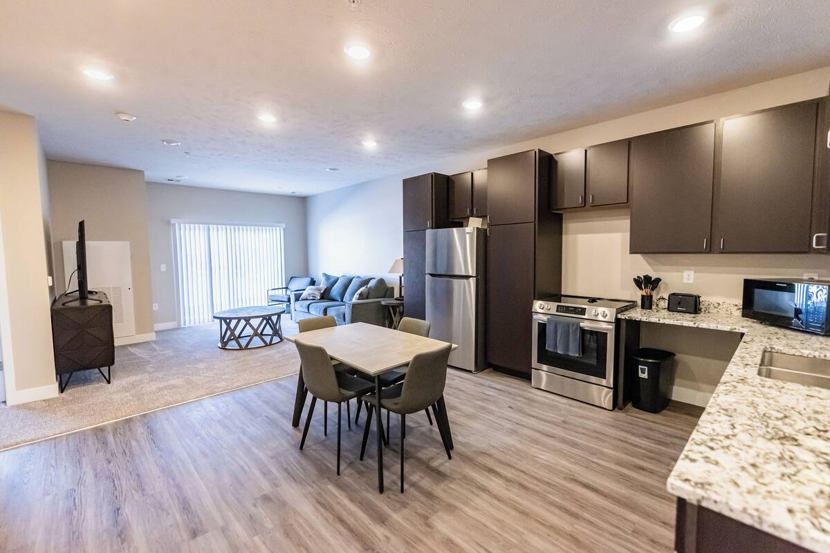 A modern kitchen with dark cabinets, a stainless steel refrigerator, a microwave, and a built-in oven. There is a dining area with a table and chairs.