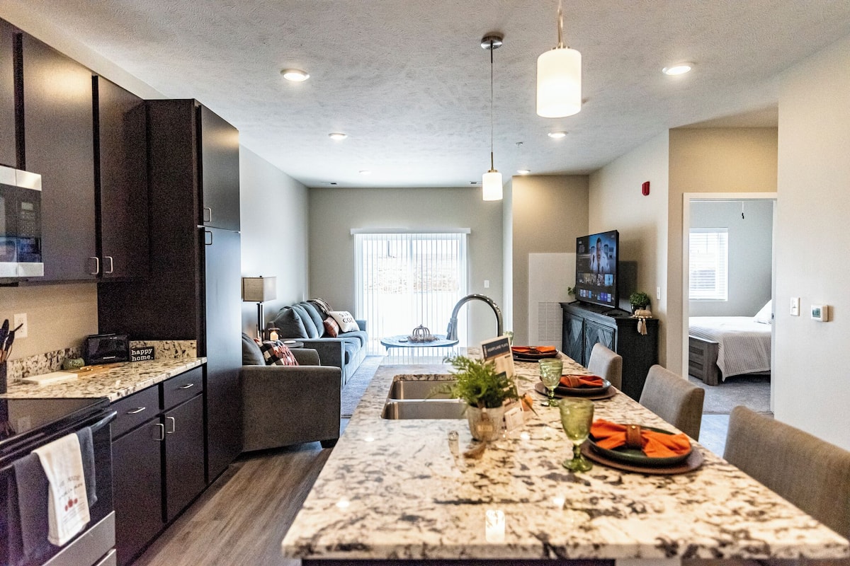 A modern kitchen with dark cabinets, a marble countertop, and a dining area with a long table.