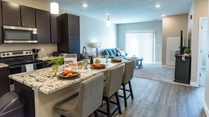 A modern kitchen with dark cabinets, a granite island, and a dining area with bar stools. - Heartland Flats  (Alliance)