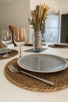 Uma mesa de jantar posta com pratos, talheres e copos de vinho, com um vaso de flores secas ao fundo.