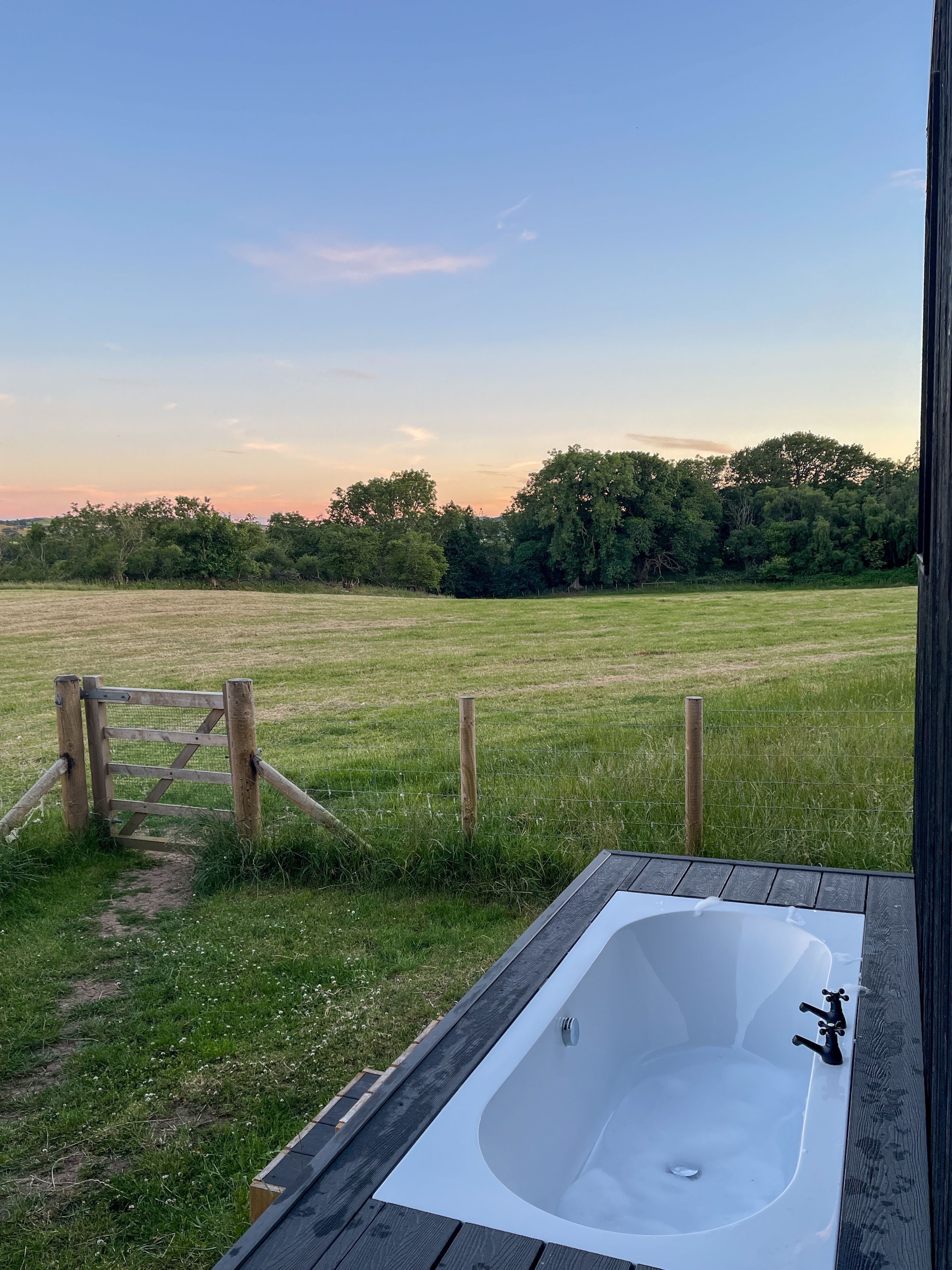 A wooden deck with a hot tub and a view of a grassy field and trees.