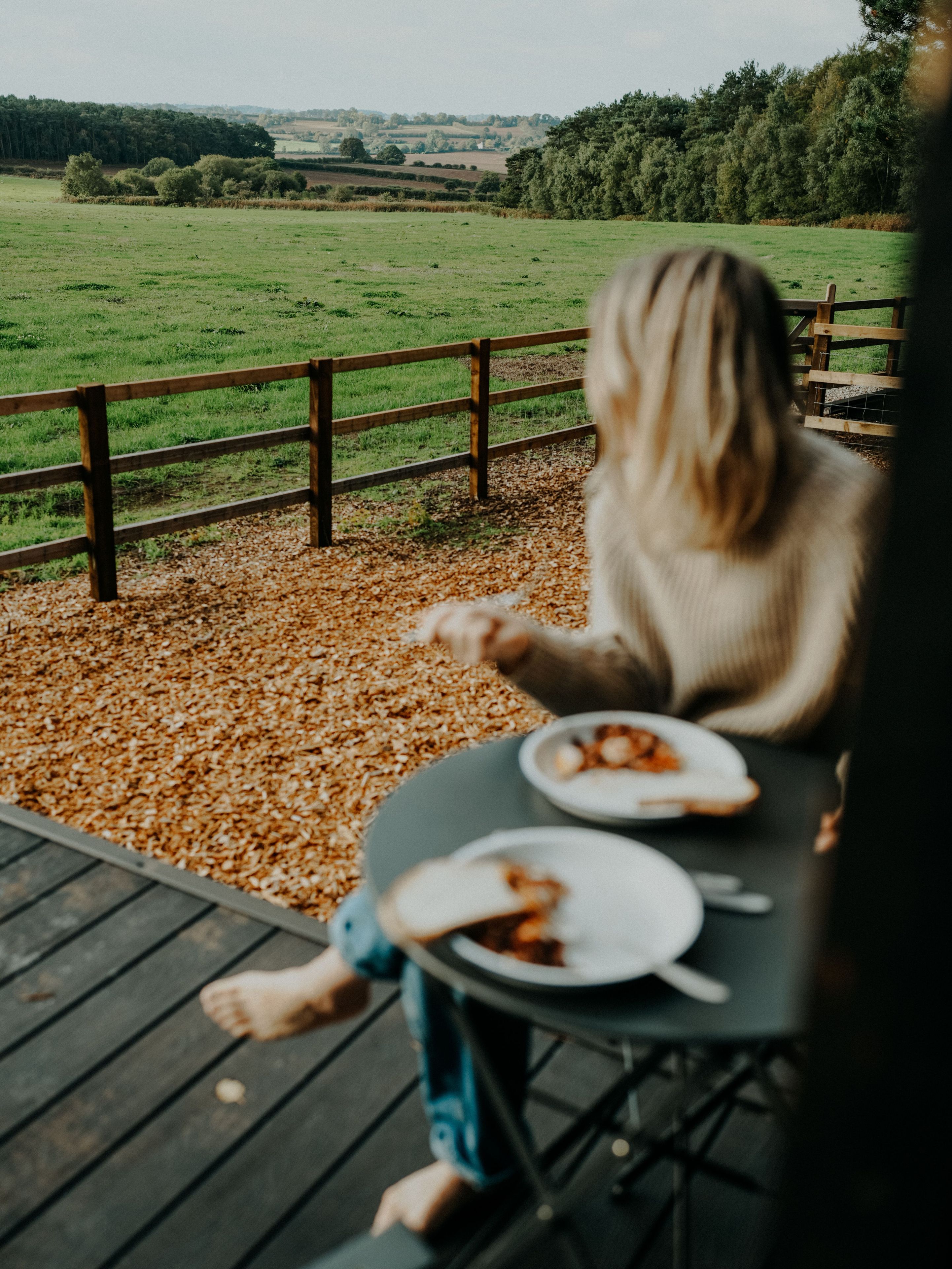 Eine Person sitzt an einem Tisch mit Essensplatten auf einer Terrasse mit Blick auf ein grünes Feld und Bäume.