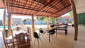 En udendørs overdækket terrasse med træborde og stole, en pool i baggrunden og en hængekøje.