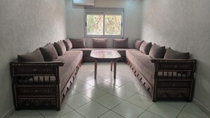 A room with a large L-shaped sofa set, a small round table, and a window with decorative iron bars. - STANDING SAIDIA APARTMENT (Saïdia)