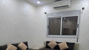 A modern living room with a sectional sofa, a coffee table, and a window with blinds. - STANDING SAIDIA APARTMENT (Saïdia)