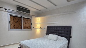 A bedroom with a large bed, a headboard, a window with decorative blinds, recessed ceiling lights, and a tiled floor. - STANDING SAIDIA APARTMENT (Saïdia)