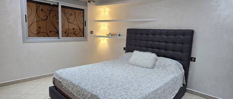 A bedroom with a large bed, a headboard, a window with decorative blinds, recessed ceiling lights, and a tiled floor.