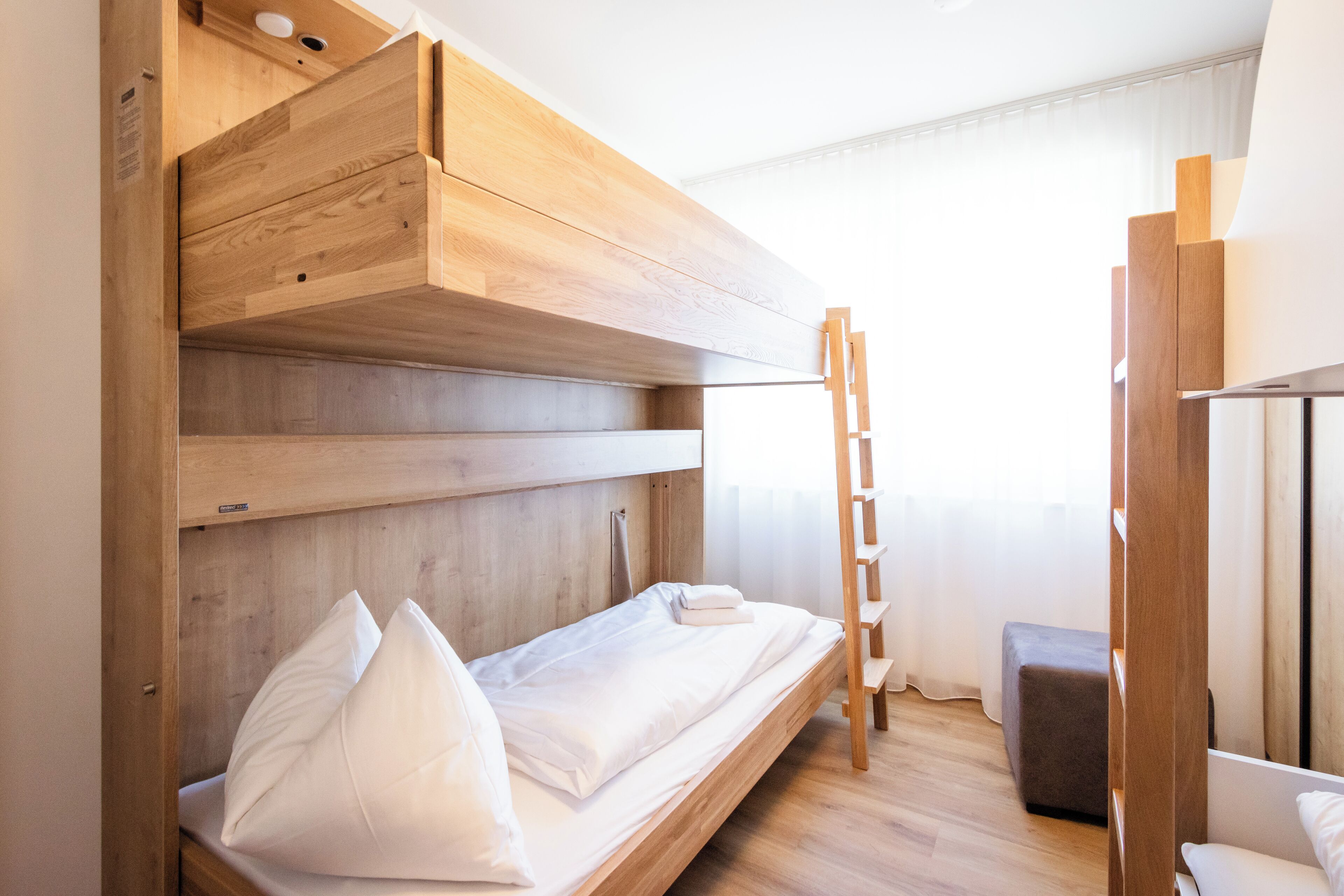 Een stapelbed met houten hoofdbord en lades, een ladder en een klein pouf in een kamer met houten vloer en een raam met gordijnen.