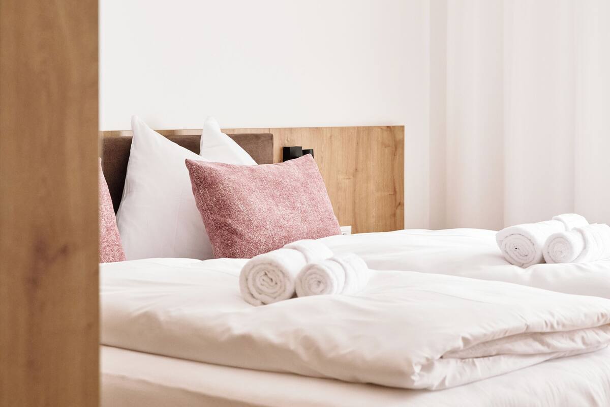 A neatly made bed with white and pink linens, a wooden headboard, and rolled towels.