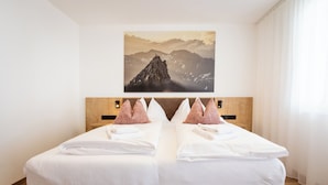 Una cama doble con sábanas blancas y almohadas decorativas rosas, un cabecero de madera y un cuadro enmarcado con un paisaje montañoso encima.