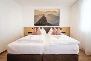 Una cama doble con sábanas blancas y almohadas decorativas rosas, un cabecero de madera y un cuadro enmarcado con un paisaje montañoso encima.