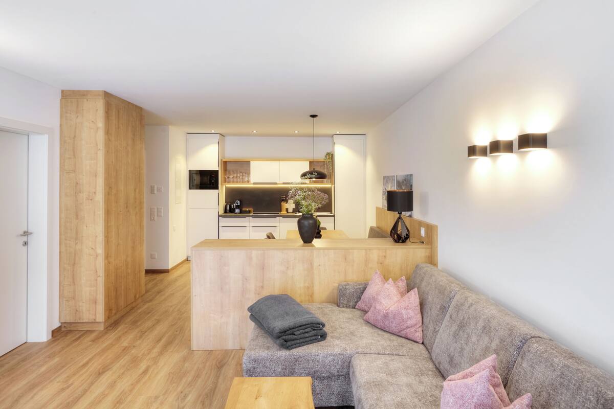 A modern living room with a grey sofa, wooden flooring, and a kitchen in the background.