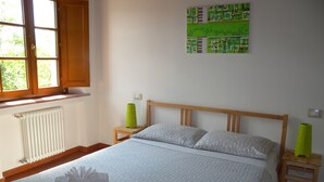 Una cama bien hecha con ropa de cama estampada, una ventana con contraventanas de madera y un cuadro abstracto verde en la pared.