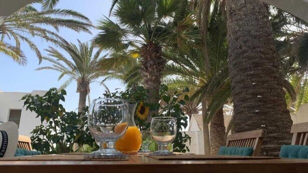 Una mesa con un vaso de jugo de naranja y un vaso de vino blanco, con palmeras y un edificio de fondo.