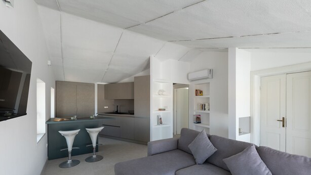 Ein modernes Wohnzimmer mit einem grauen Sofa, zwei weißen Barhocker und einer Küche im Hintergrund.