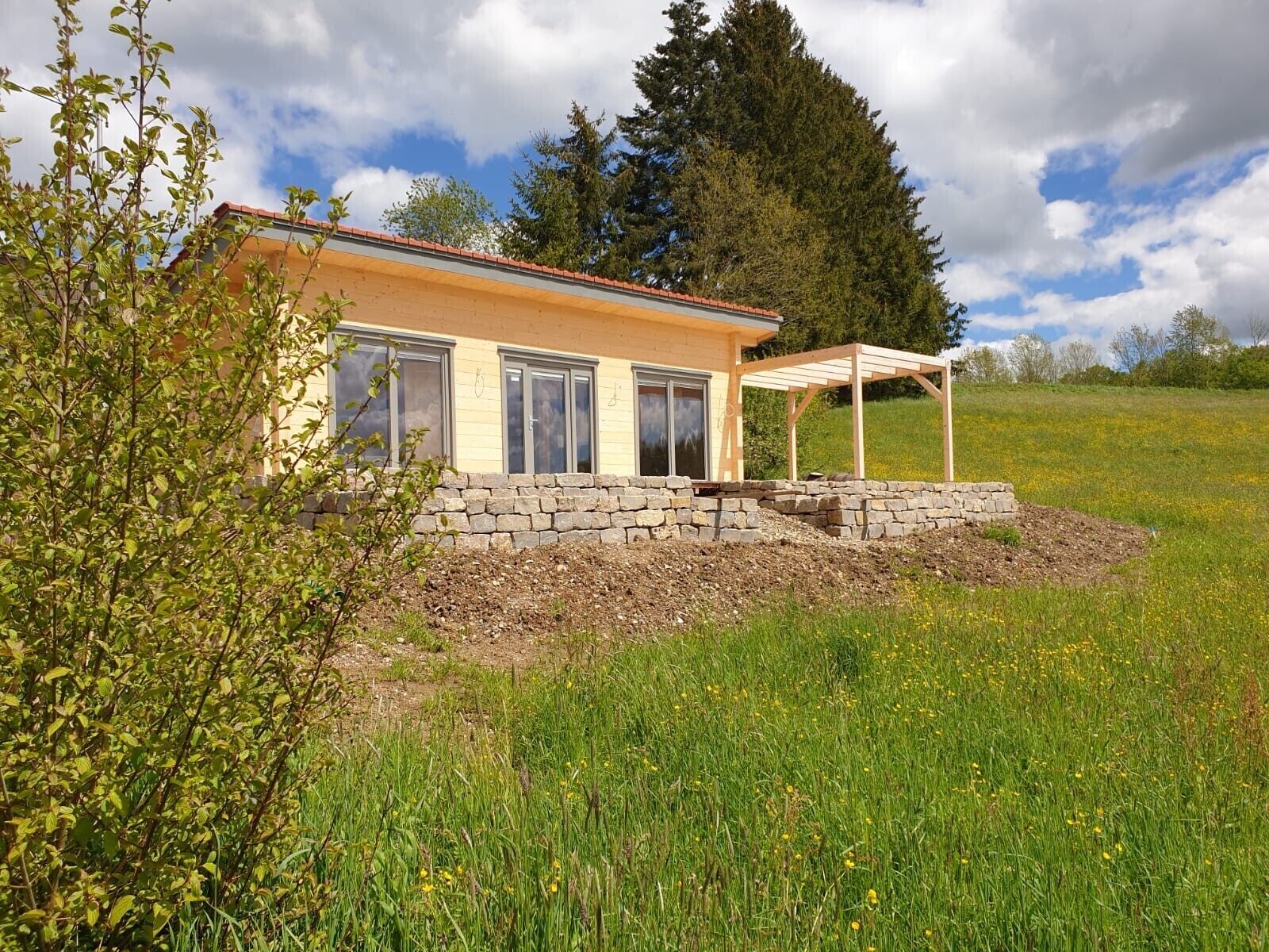 A house with a stone foundation, a wooden pergola, and a grassy field.