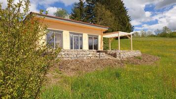 A house with a stone foundation, a wooden pergola, and a grassy field.