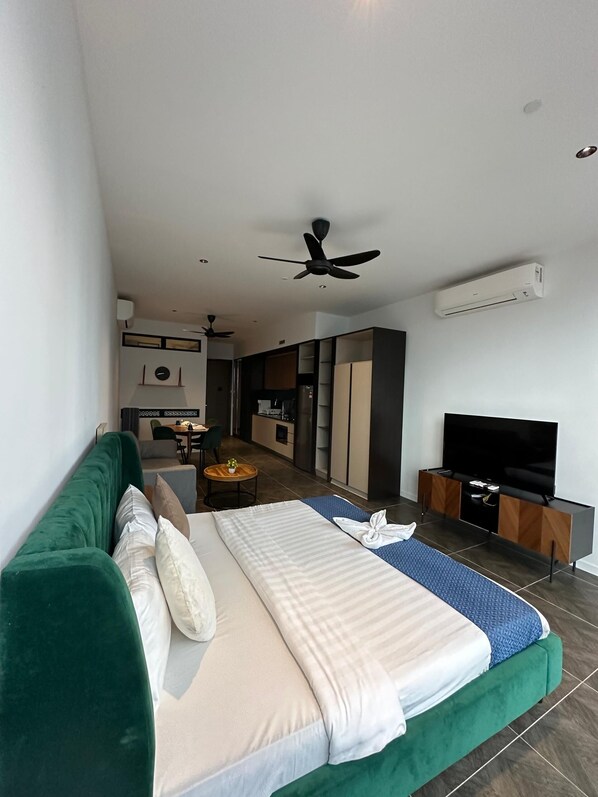 Una habitación de hotel moderna con un sofá verde, una cama con colcha azul y blanca, un televisor sobre un soporte de madera y un ventilador de techo.
