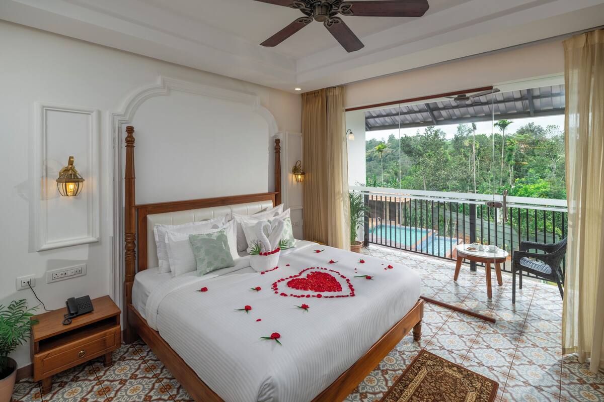 A hotel room with a bed, a nightstand, a telephone, a ceiling fan, a balcony with a view of a pool and greenery.