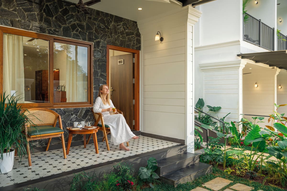 A woman sitting on a chair outside a building with a stone wall, a wooden door, and a balcony with railings.