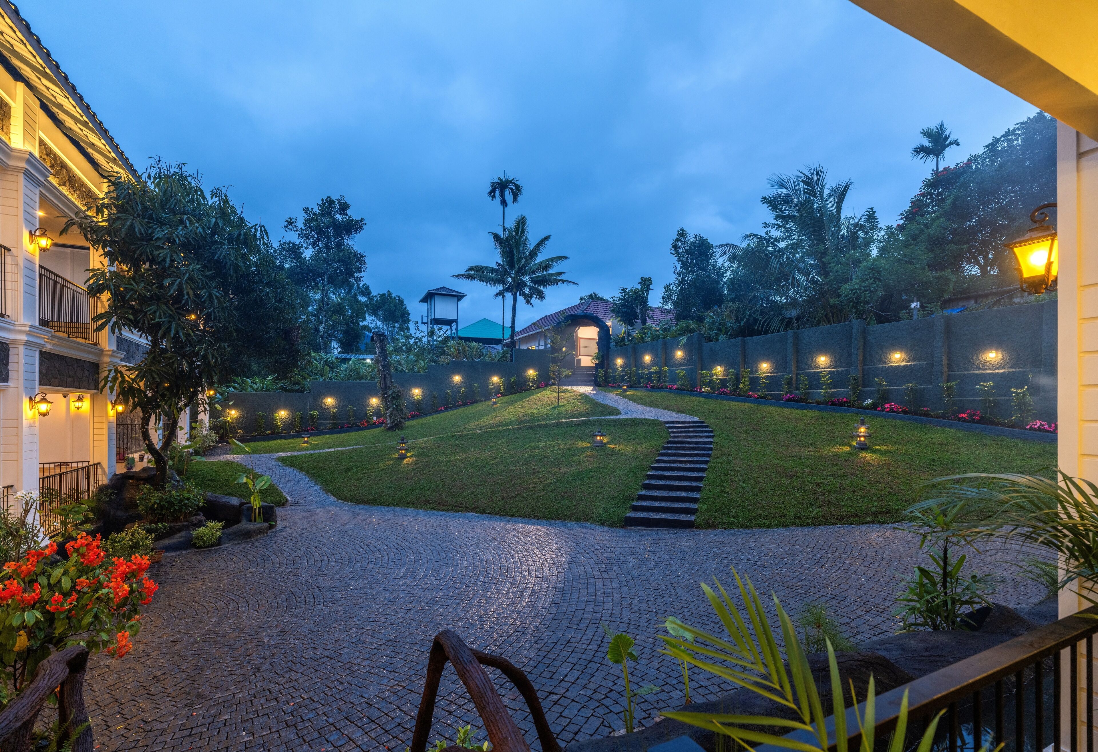 A well-lit courtyard with a paved pathway leading to a building entrance, surrounded by lush greenery and palm trees.
