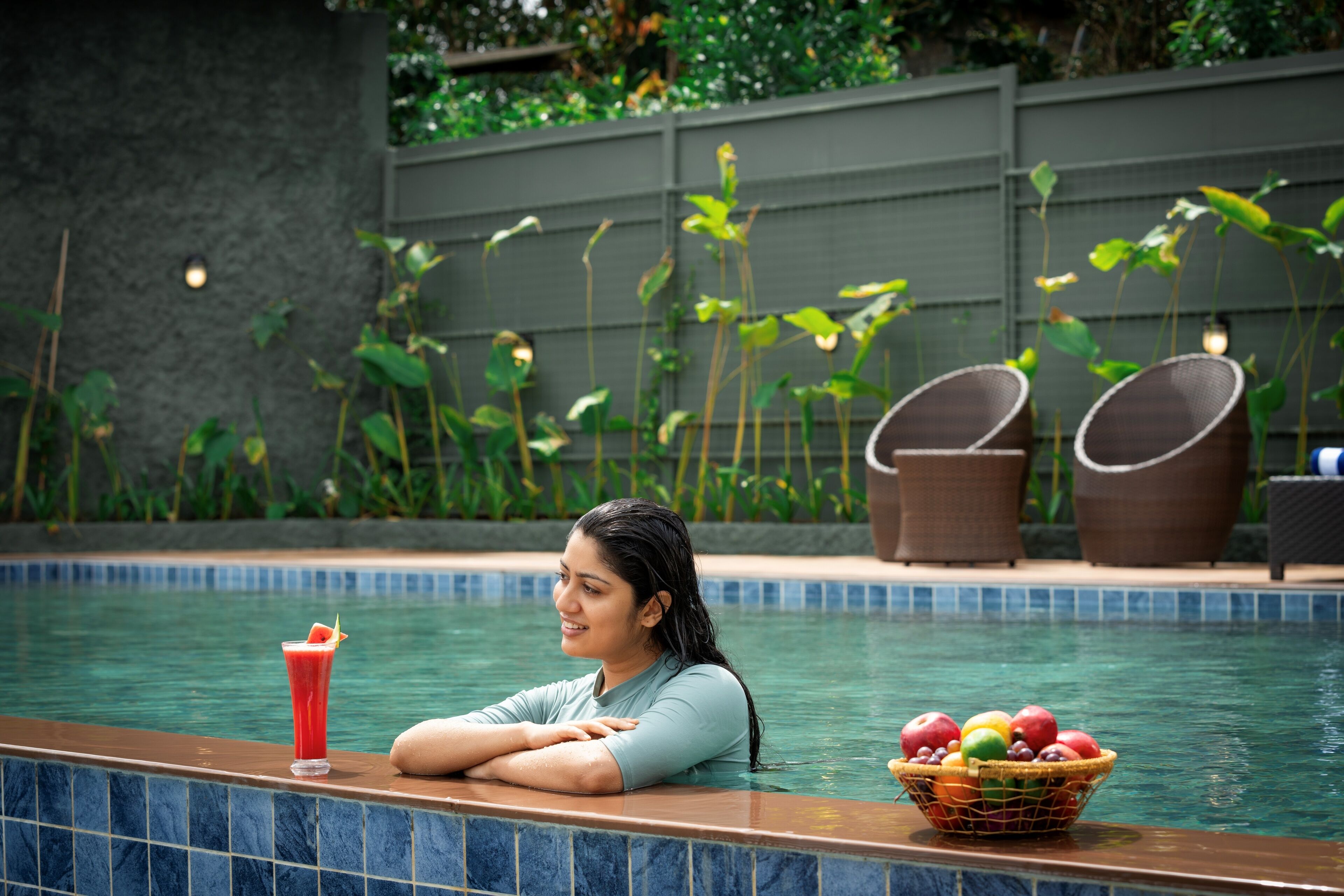 A poolside area with a person leaning on the edge, a drink, and a fruit basket.