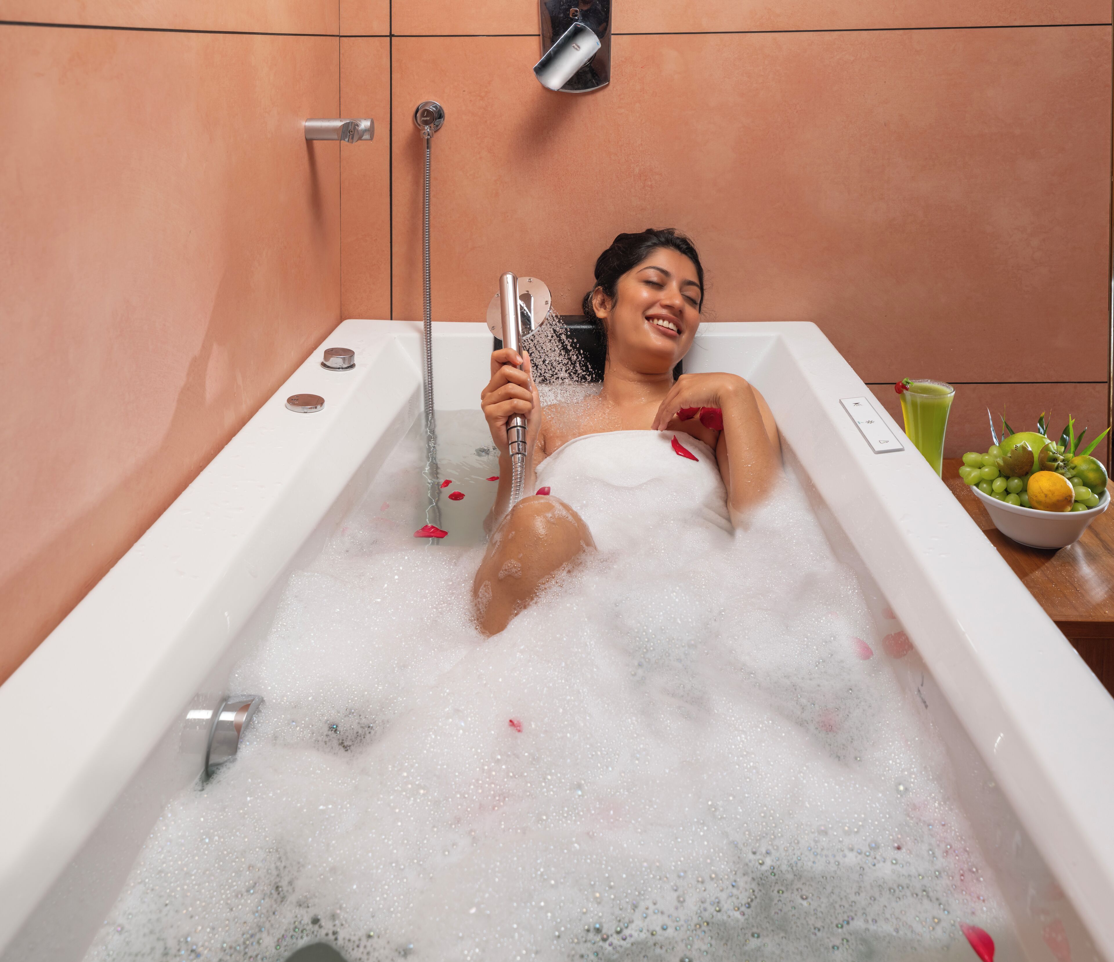 A person relaxing in a bathtub filled with bubbles, holding a showerhead. There is a bowl of fruit and a glass of green drink on a wooden table beside the tub.