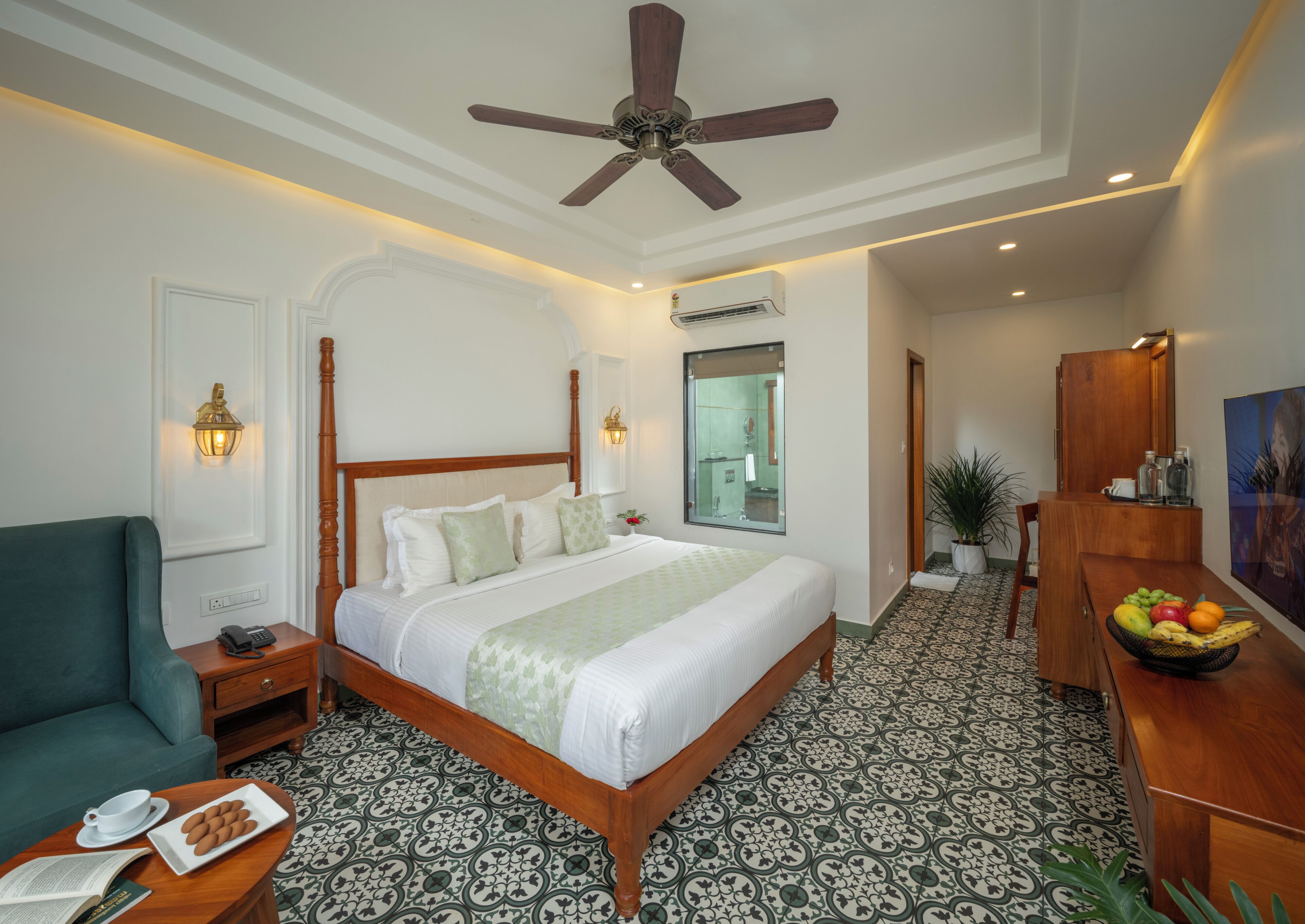 A hotel room with a large bed, a desk with a fruit bowl, a green armchair, a ceiling fan, and patterned floor tiles.
