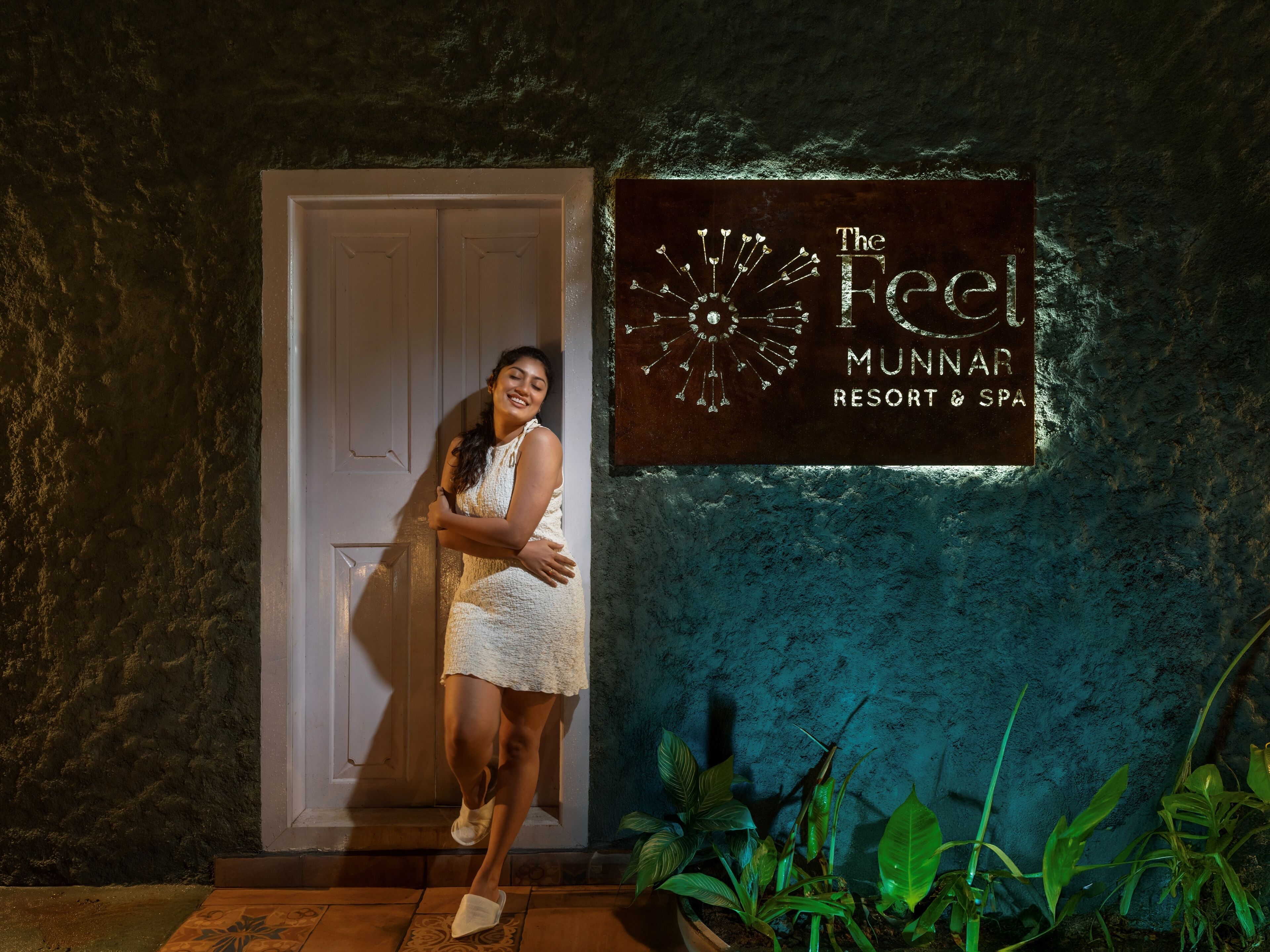 A woman in a white dress standing in front of a door with a sign that reads "The Feel Munnar Resort & Spa".