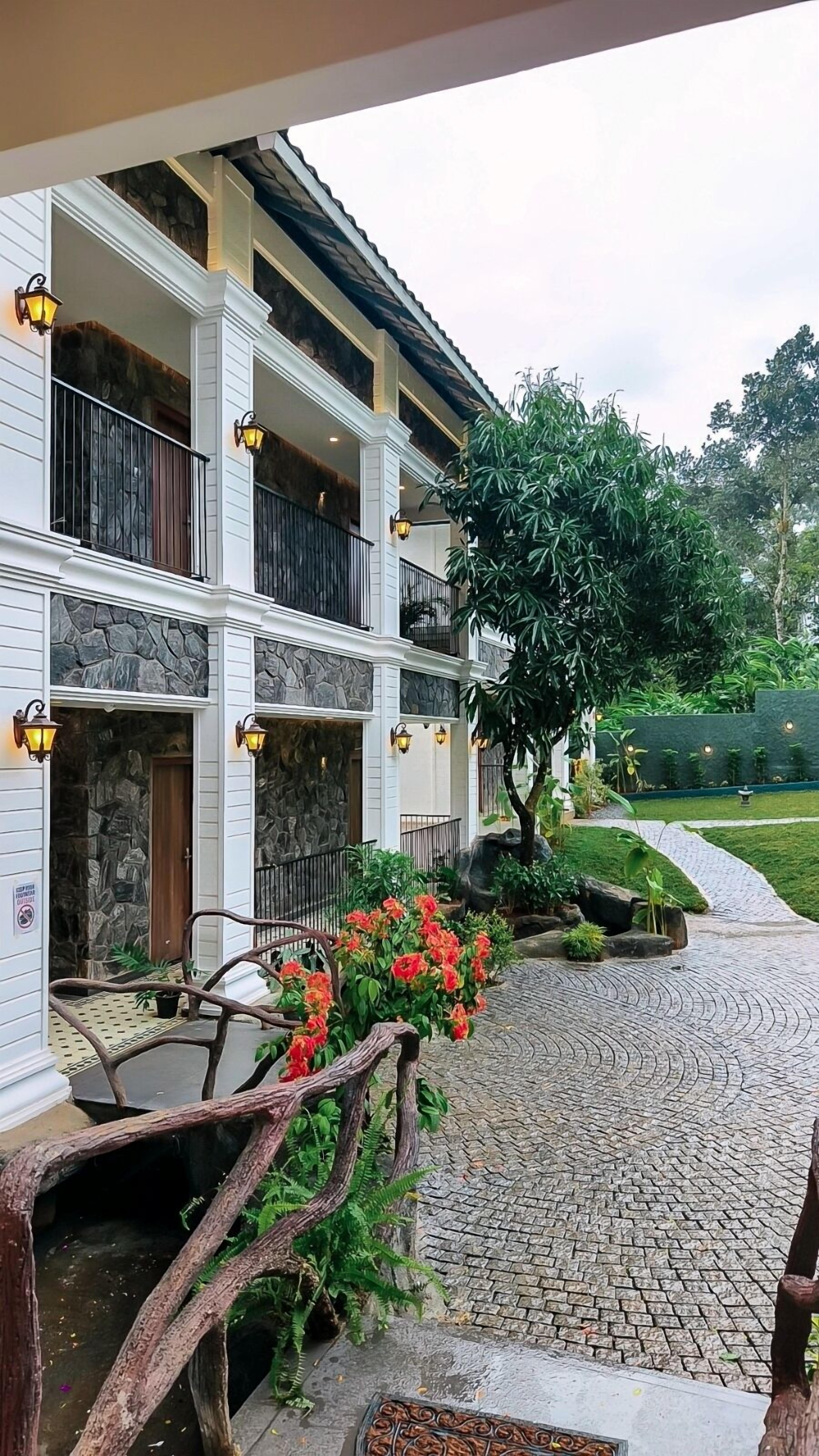 A two-story building with stone facade and balconies, a paved pathway, and a small garden with red flowers.