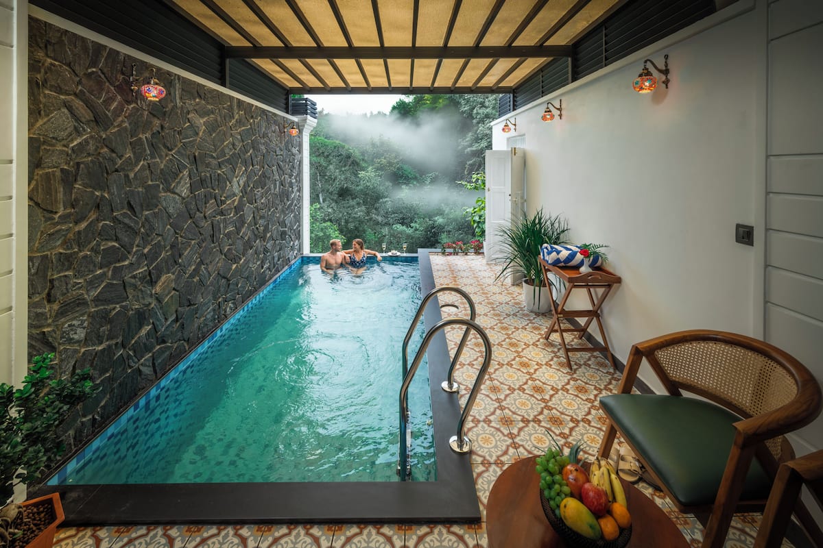 A stone-walled infinity pool with a clear view of the surrounding greenery, a wooden deck, and a seating area with a fruit bowl.