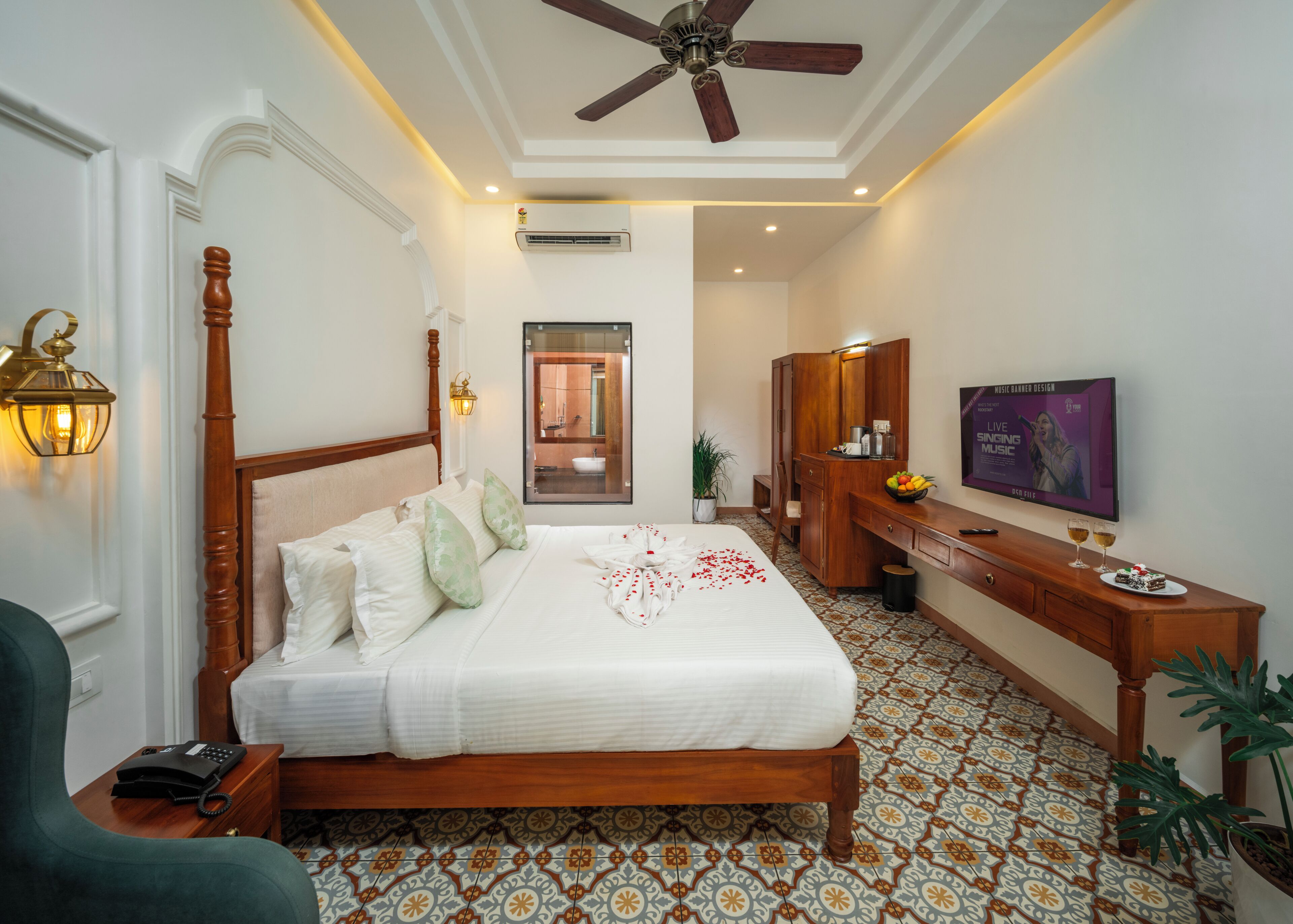 A hotel room with a wooden bed, a desk with a flat-screen TV, a ceiling fan, a bathroom mirror, and a patterned floor.