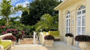 Une terrasse ensoleillée avec un canapé blanc, des plantes en pot et un portail blanc donnant sur un jardin luxuriant.