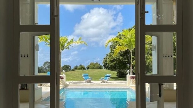 Une zone de piscine avec deux chaises longues et des palmiers, vue à travers une porte vitrée.
