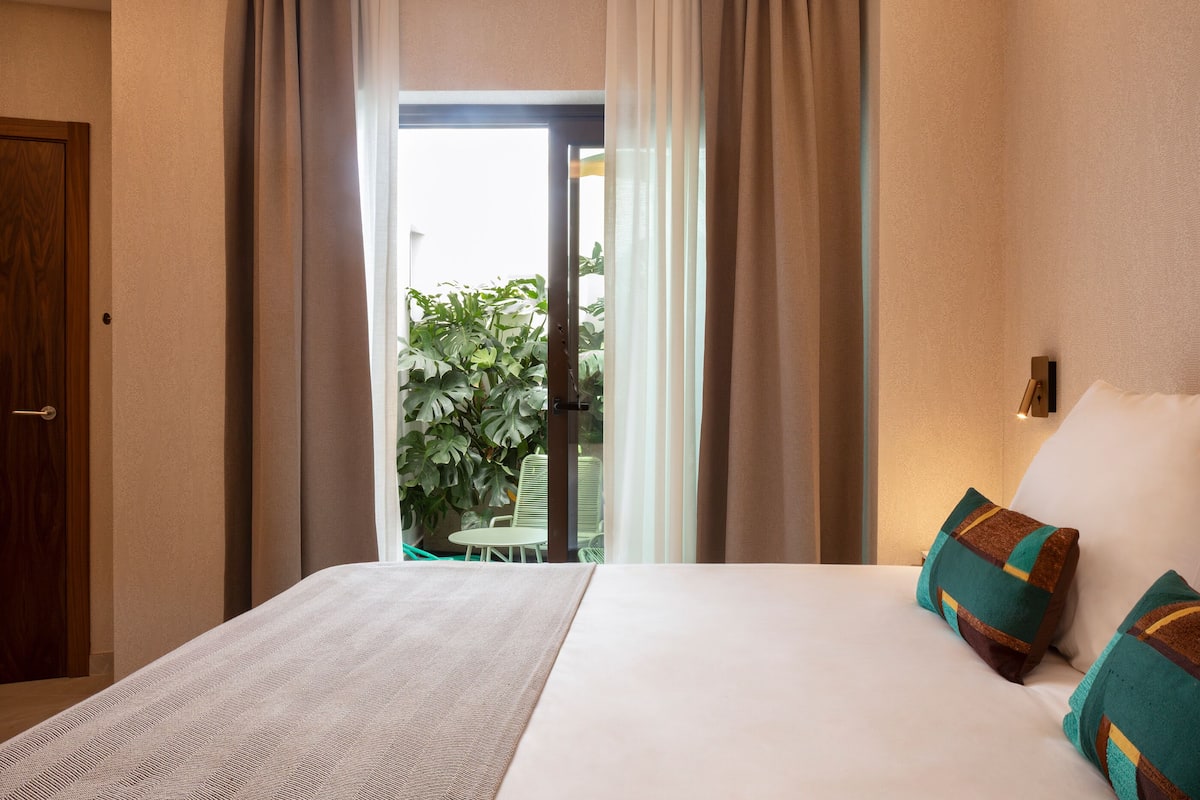 A hotel room with a bed, curtains, a view of greenery outside, and a door.