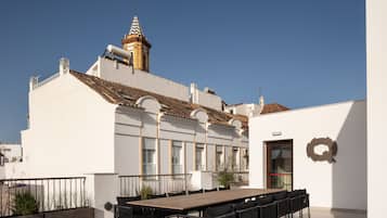A rooftop terrace with a long wooden table and black chairs, a white building with a tiled roof, and a clear blue sky.