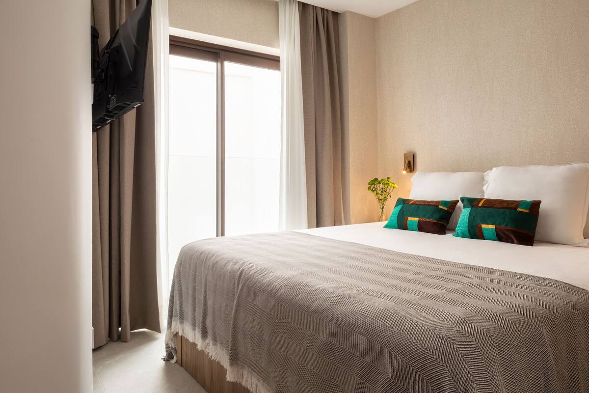 A hotel room with a bed, curtains, a flat-screen TV mounted on the wall, and a window with curtains.