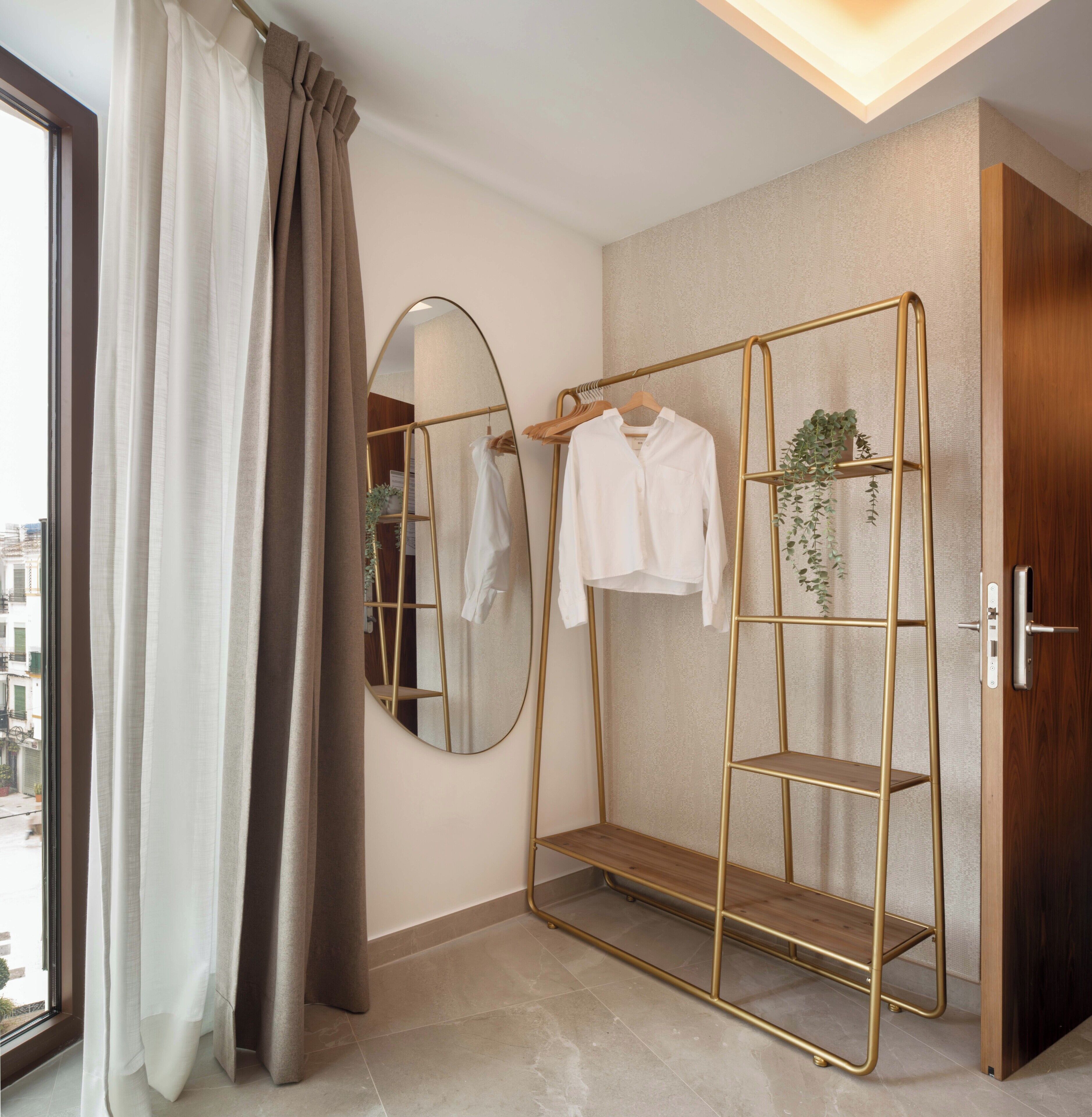 Een kamer met een groot raam, een spiegel, een gouden kast en een hangplank met kleding en een plant.