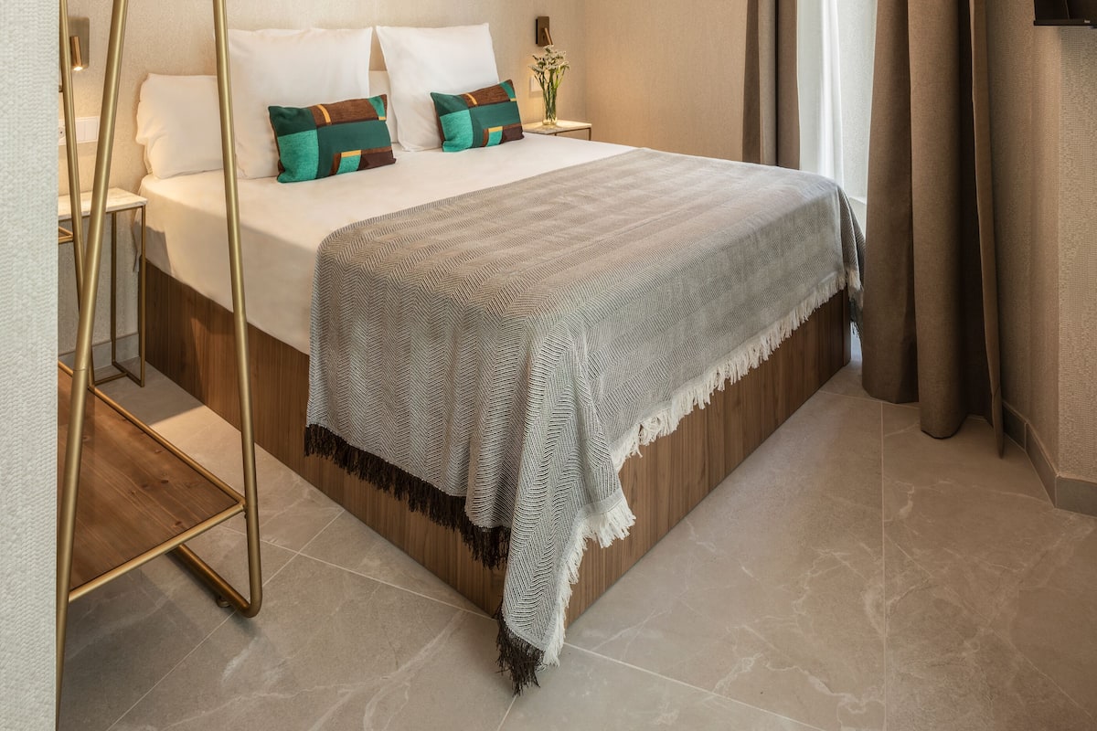 A bedroom with a bed, bedside tables, a hanging light, a mirror, and a view of the outside.