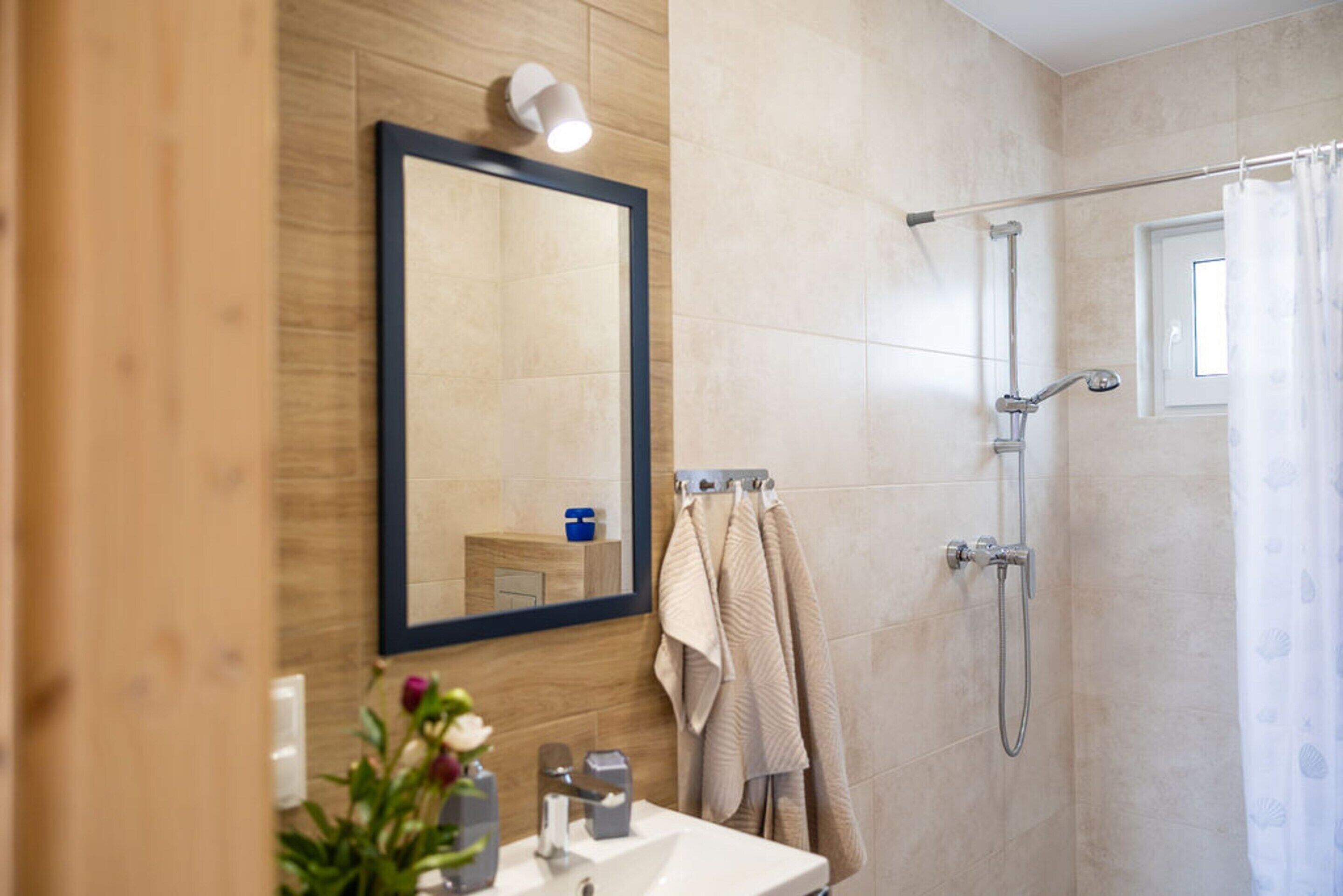 Een moderne badkamer met een houten wastafelmeubel, een grote spiegel, een douche met handdouche en een raam met gordijn.