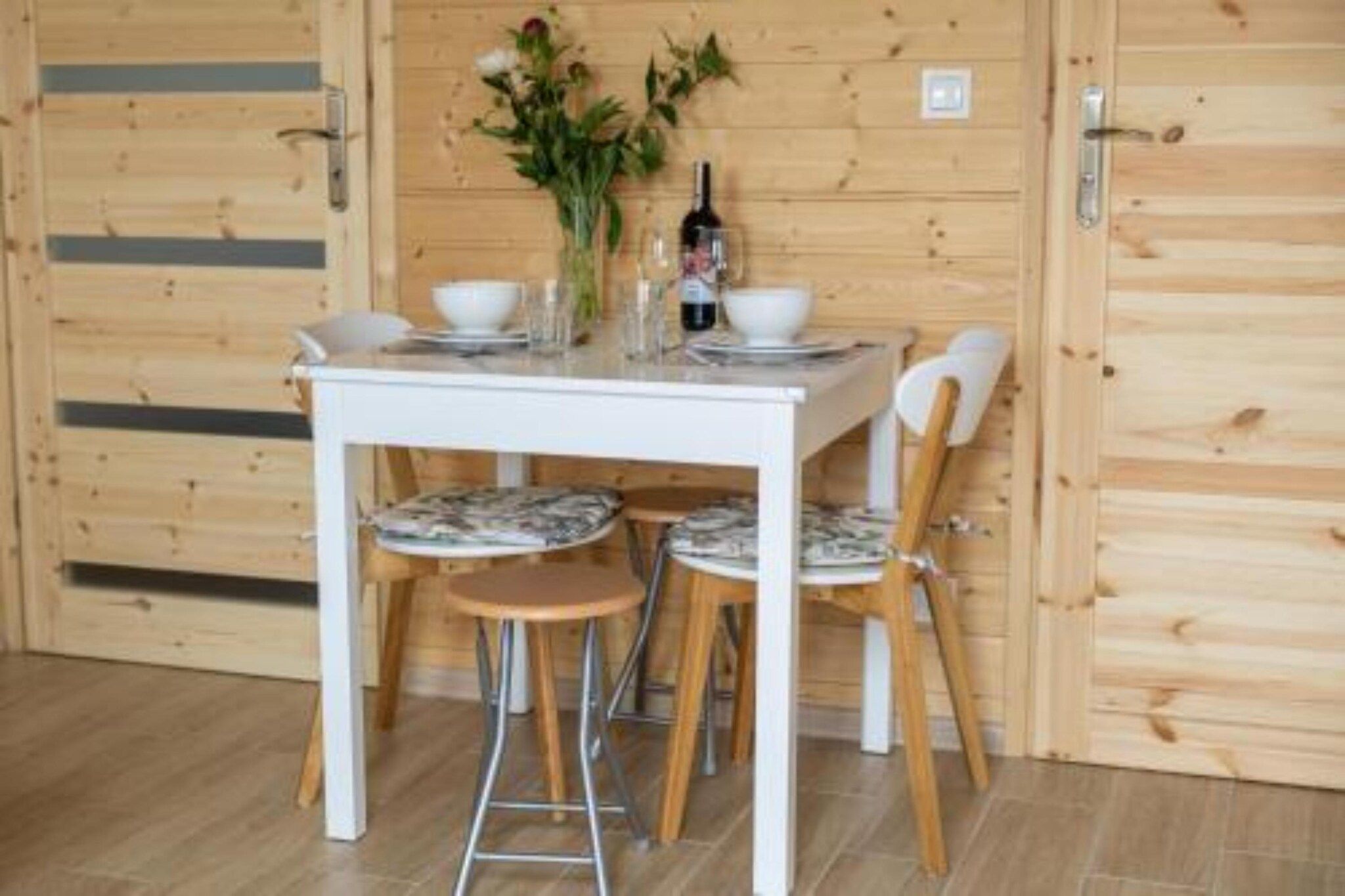Un intérieur en bois avec une table et des chaises blanches, un vase rempli de fleurs et une bouteille.
