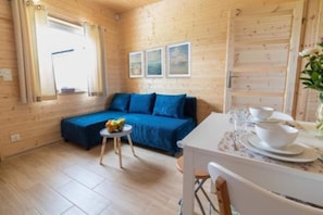 A cozy cabin interior with a wooden floor, a blue sofa, a small table with a fruit bowl, and a dining table set for two.